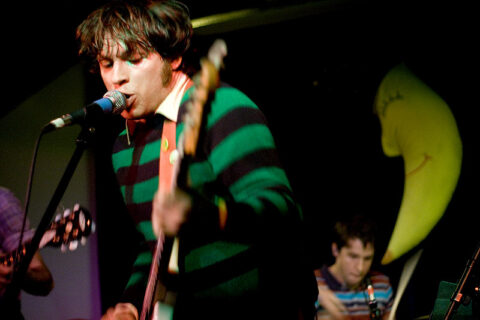 Man in striped long sleeve shirt singing into a microphone while playing a guitar. Another man is playing drums in the background, and a third person is playing the guitar out of frame.