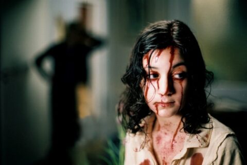 White woman with dark hair covered in blood. There is a blurry silhouette of a person in the background.