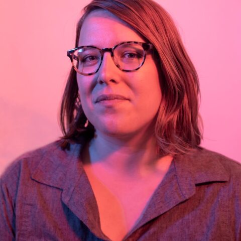 A portrait headshot of Kelsey in pink light. They are a white woman with light brown shoulder length hair, round glasses, no makeup, and a slight smile.