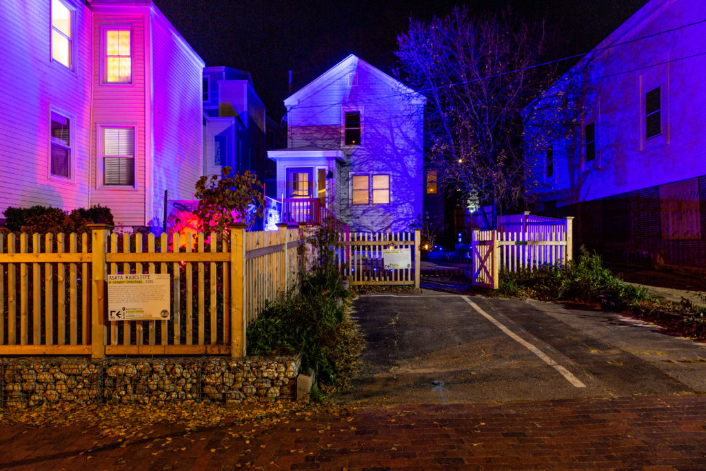 nighttime exterior photo of a house and yard with wooden fence and pink and purple lighting
