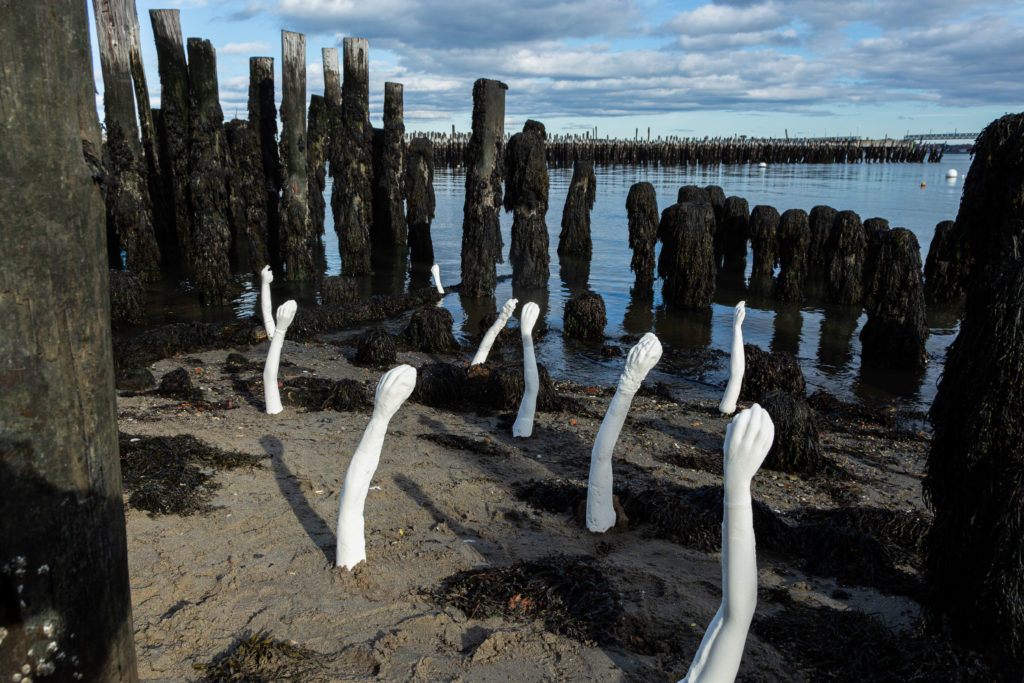 photo of a sandy beach with old wooden pillars and sculptures of arms made of sugar protruding from the san
