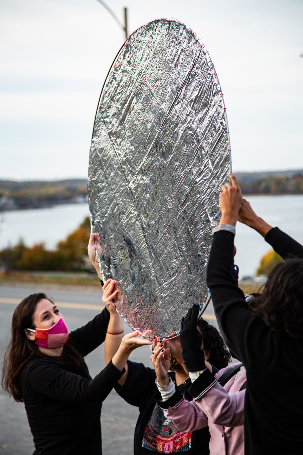 exterior photo of a small group of people holding up a large silver disc, with ocean in the background