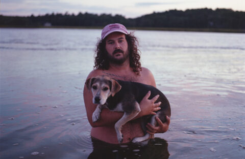 It's Colby! He's waist-deep in the ocean and holding his dog Tina. It's a beautifully Maine picture and weirdly intimate. Colby is wearing a hat. It seems to be near dusk. A light sunset cascades across the water. What a day.