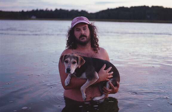 It's Colby! He's waist-deep in the ocean and holding his dog Tina. It's a beautifully Maine picture and weirdly intimate. Colby is wearing a hat. It seems to be near dusk. A light sunset cascades across the water. What a day.