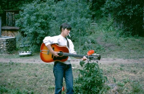 A woman in jeans stands alone near the woods, playing a guitar and smoking a cigarette.