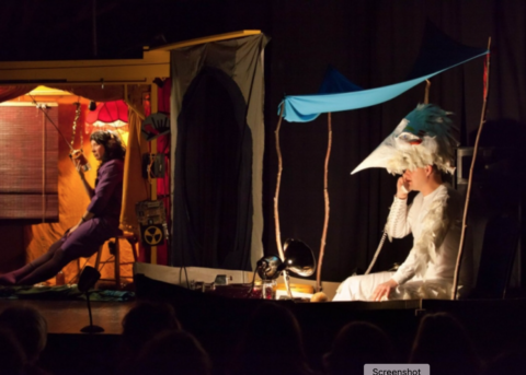 A photo from the play Below and Beyond, a figure seems to be in a white bird costume talking on the phone, and a second figure is in silhouette on a walk talky in the background.