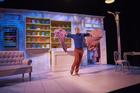 set of a play featuring a man dancing in front of colorful shop shelves