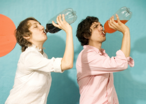 The two members of The Blow chugging what appears to be a bottle of cola and a bottle of orange soda against a teal backdrop.