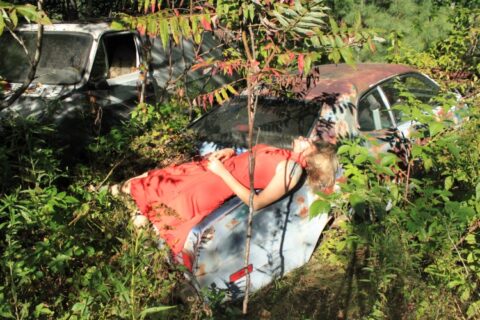 Digital print of a girl in an orange dress laying across a car trunk in a wooded area