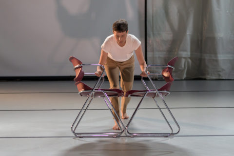 A photograph of Lailye Weidman performing a works in progress showing of the work, bringing two chairs together in a white t-shirt and khaki pants.