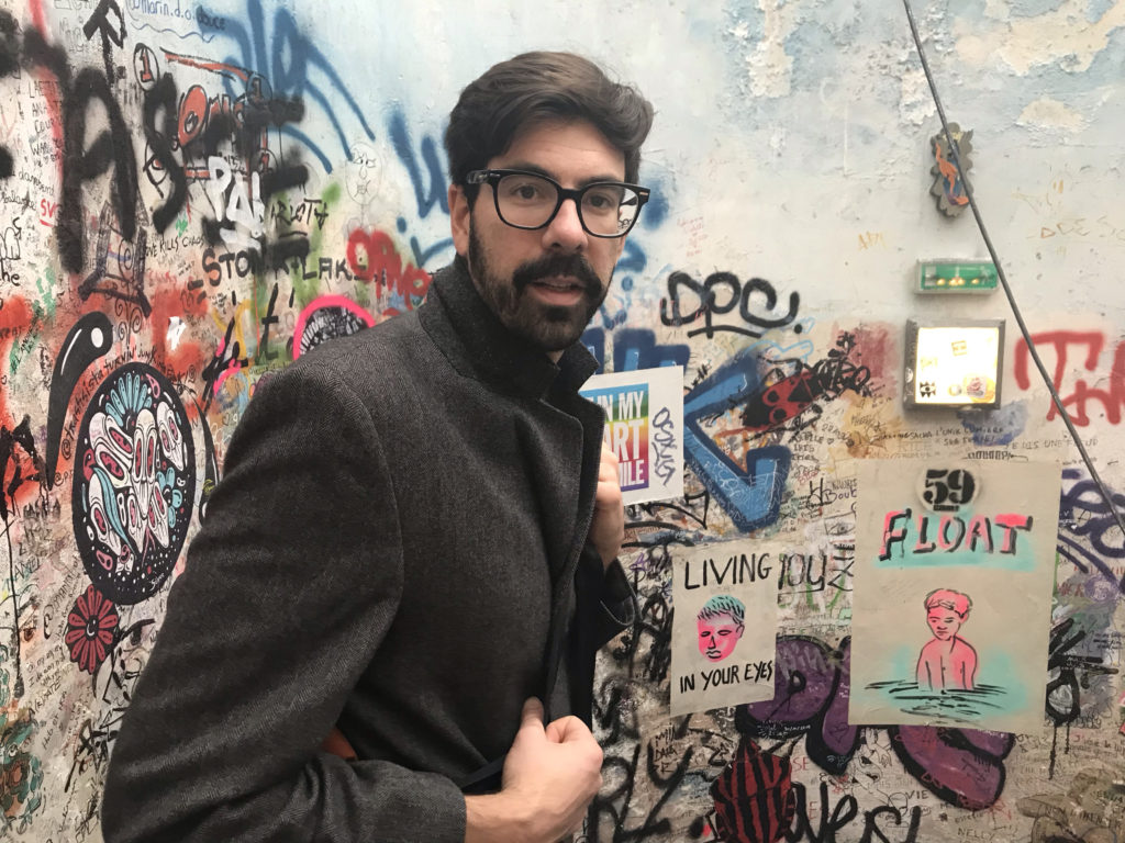 A photograph of Anthony Moffa in glasses with a black coat against a graffitied background.