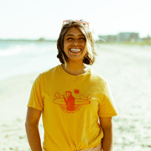 Pam Chevez on a beach smiling with orange sunglasses above her head and a yellow surfing shirt.