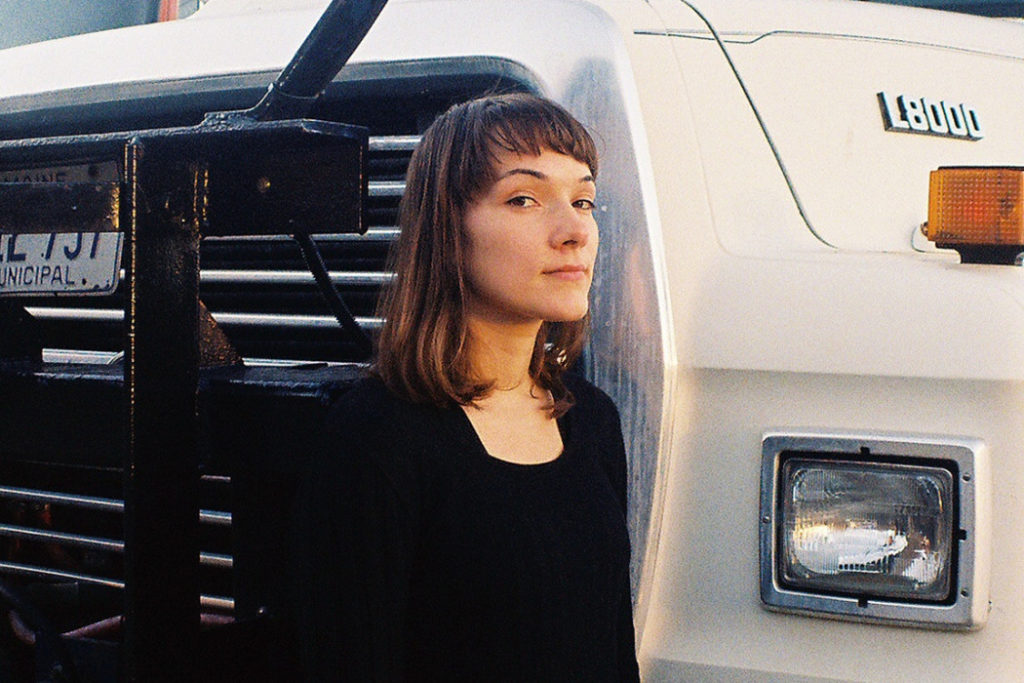 Lisa/Liza beside a white truck in a black shirt and sweater looking with a slight side-eye expressionlessly into the camera. 