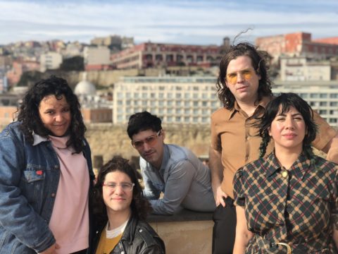 A Portrait of the five members of Downtown Boys on a rooftop with a cityscape background.