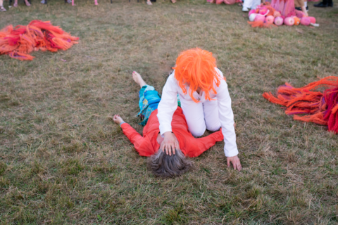 An image of a bright orange wigged figure with her hand to the forehead of a figure lying on the grass.