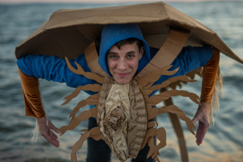 A photograph of artist Eli Nixon in a blue hoodie wearing a cardboard sculpture of a horse shoe grab form, against a backdrop of the ocean.