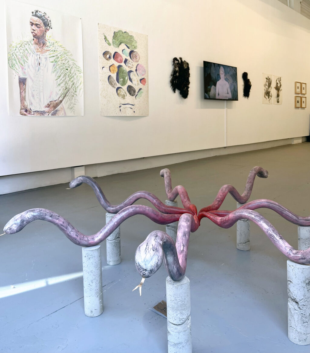 A snake sculpture by Brian Smith with a wall of 2D works and a monitor in the background, an installation view of the gallery.