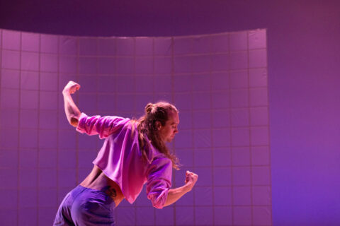 A photograph of dancer Tristan Koepke with long hair in a pink top and purple pants, back facing the camera looking to the right side, amid pink light.