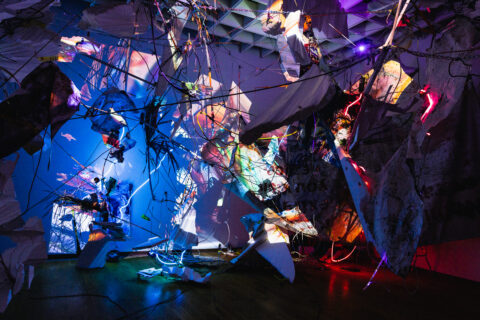 An image of an installation with projections, screens, neon, and various detritus hanging from the ceiling of OMA.