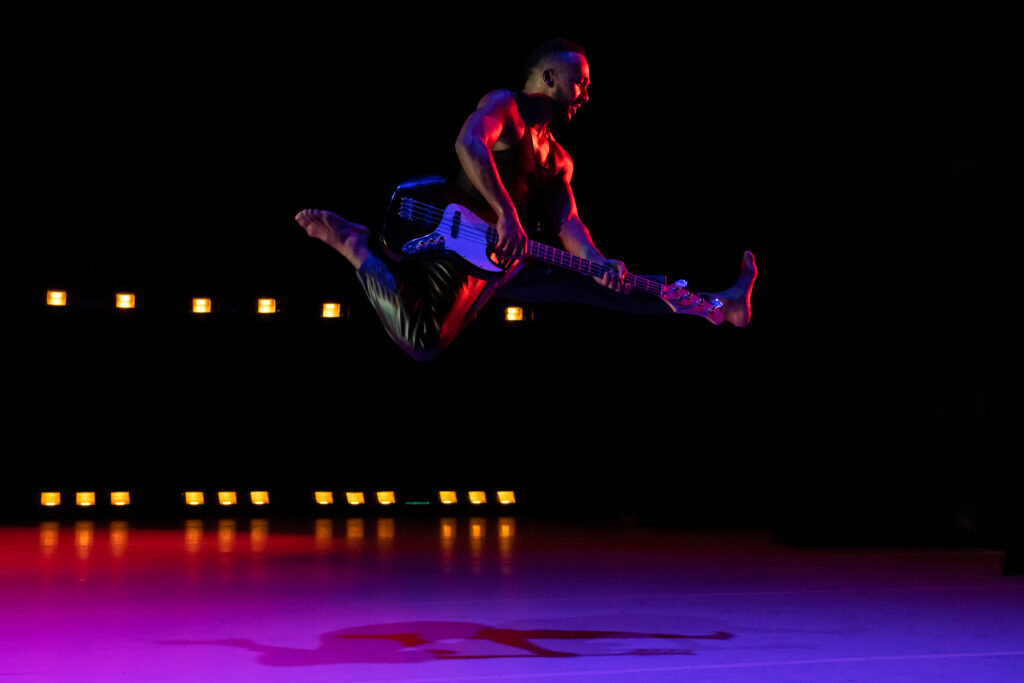 A dancer jumping with a bass guitar in hand and knees bent in red and blue lights.