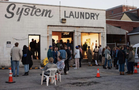 An image of the 82 Parris gallery reading System Laundry on the side of the building with the doors open and people outside.