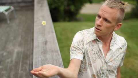 Scott McPheeters, a white man wearing a short sleeve shirt, stands with his arm outstretched and palm up while resting over a wooden plank. the image is from a Subcircle dance film, by Jorge Cousineau