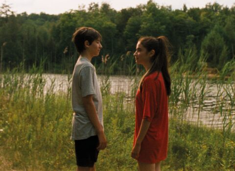 two young people staring closely at each other in front of a lake with tall grass in front of it