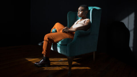 A high contrast photo of Vagabond sitting in a teal chair with orange pants and a white top.