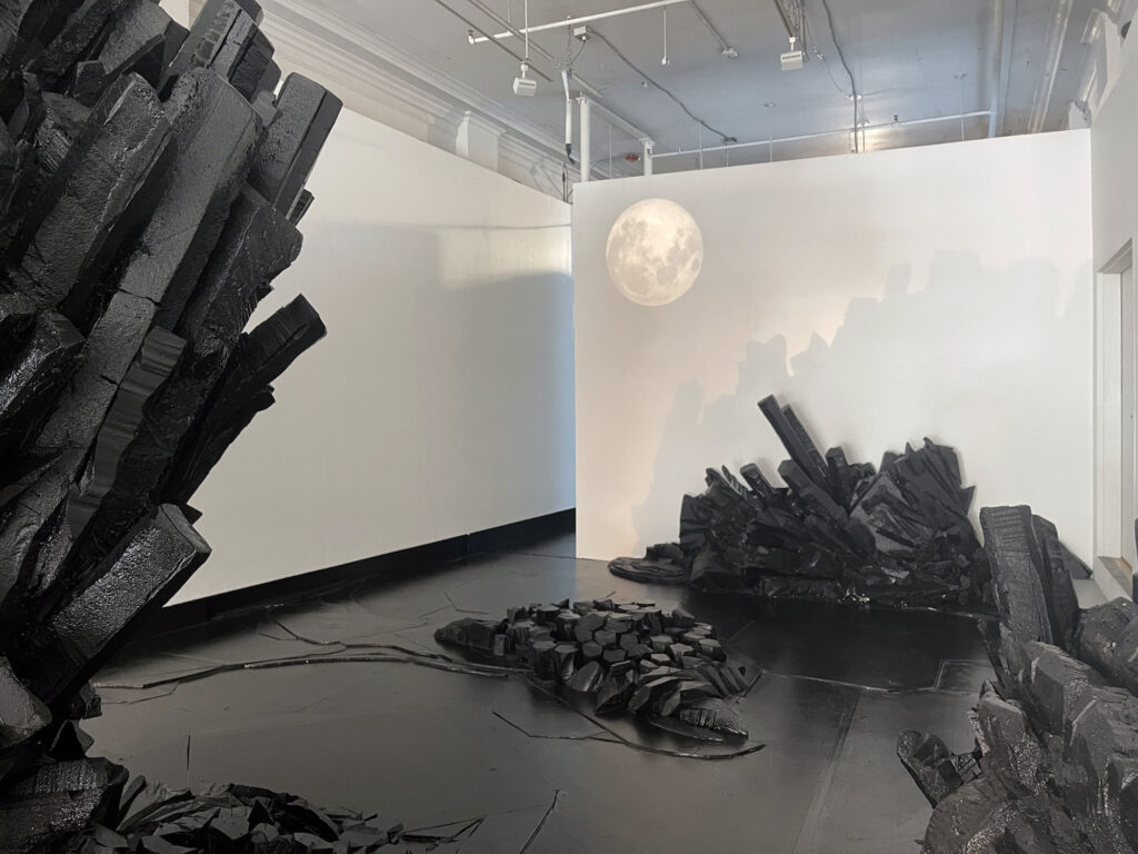 Black sculptural basalt columns surround the SPACE gallery with white walls and a projection of the moon in the corner.