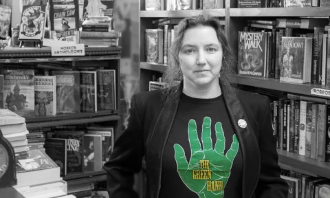 BLack and white photo of a woman standing in front of bookshelves with green hand book shop logo on shirt