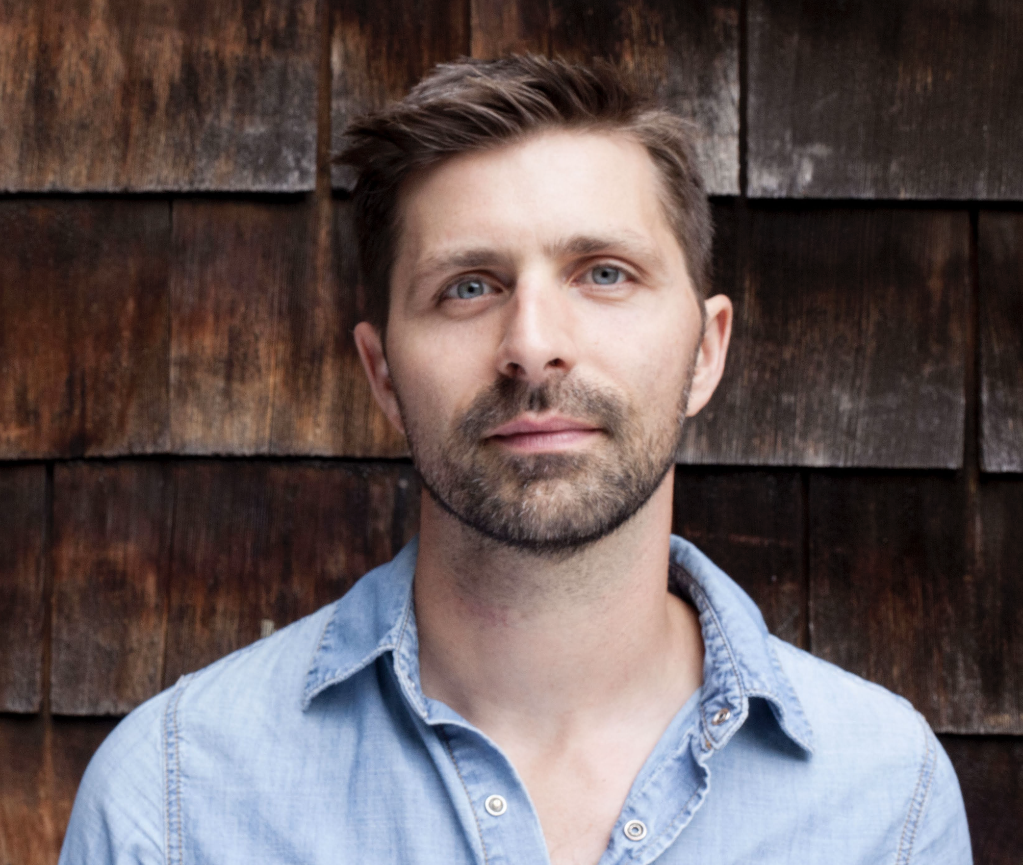 A headshot of the author in a light blue button down shirt against dark wood shingle siding. 