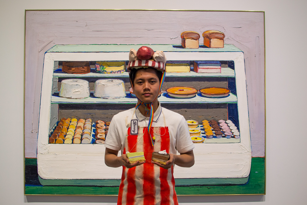 man in striped apron and ceramic hot dog hat holding two ceramic pastries standing in front of a painting of a bakery case