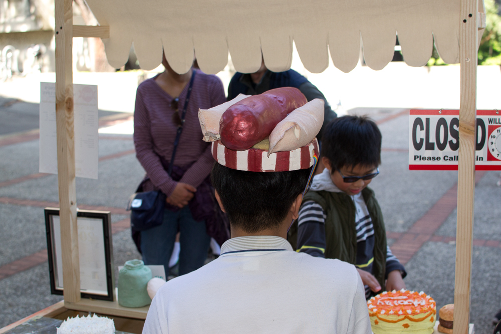 the back of a mans hea wearing a ceramic hot dog hat facing two people approaching his table