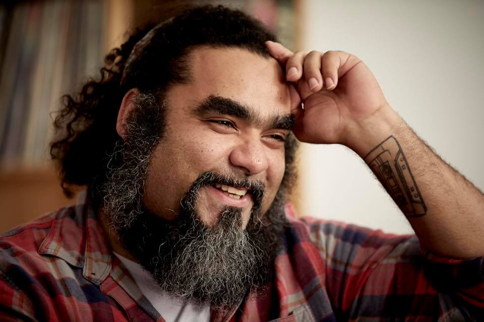 A photograph of DJ Mo Nunez (aka Mosart212) smiling and laughing, with a beard, with his hand to his face.