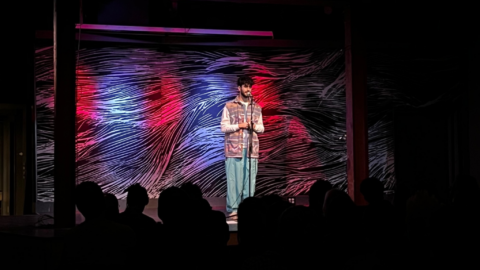 An image of a person on the SPACE stage with colored lights in the background telling a story at the microphone.