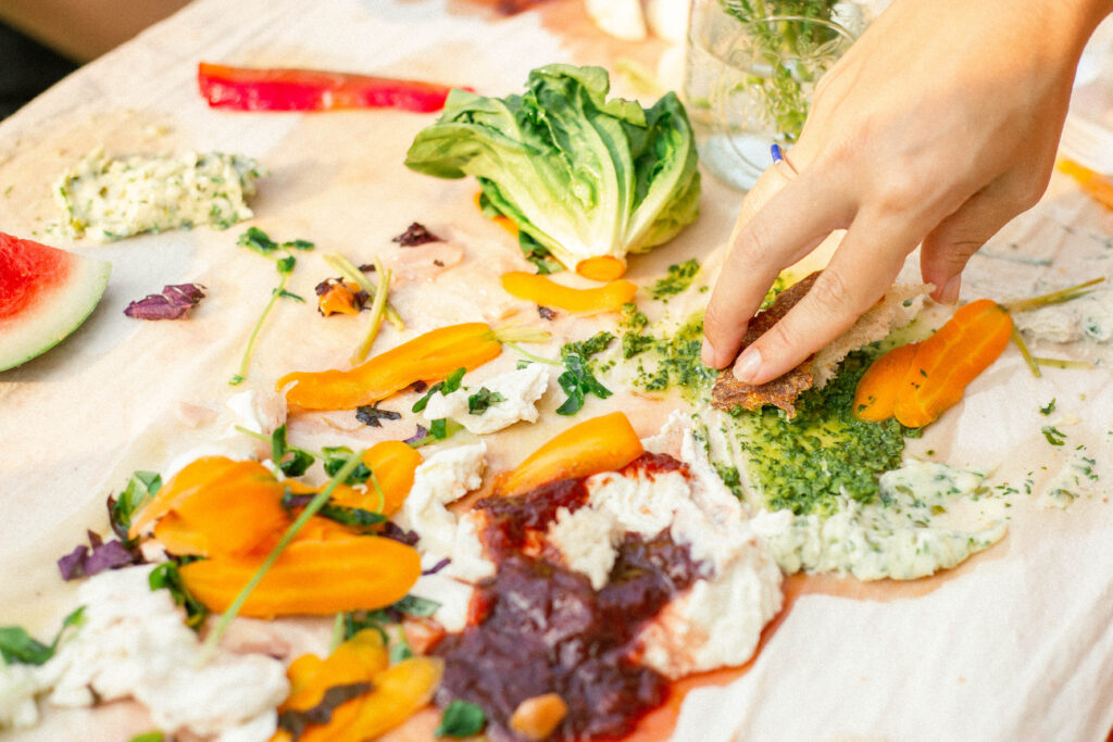 a hand reaches for a colorful spread of food against a tablecloth 