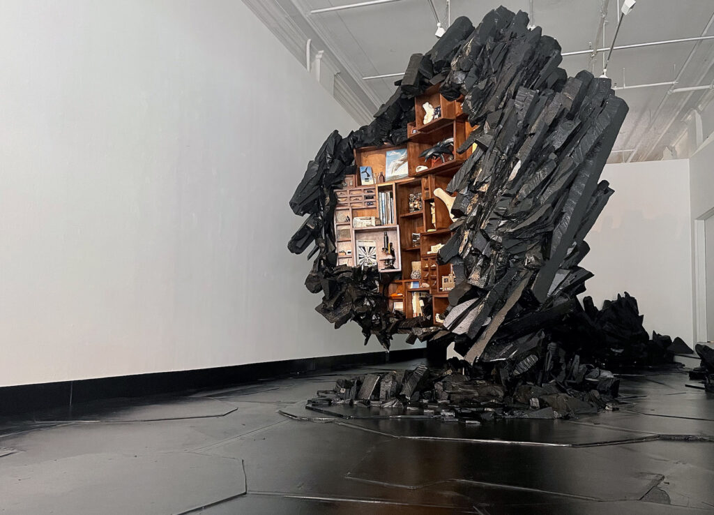 A black sculpture containing a Victorian-style cabinet of curiosities within it is in the center of a white room with a black floor. The black form looks like basalt columns by Jonathan Latiano.