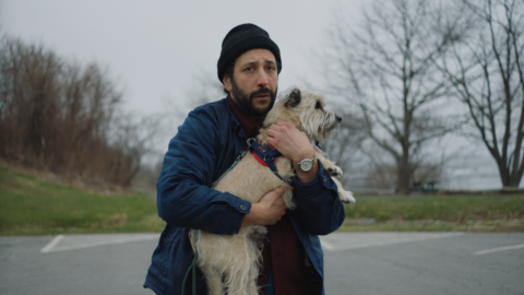 a man clutches a small fuzzy beige dog in fear, standing in a parking lot surrounded by bare trees and grass