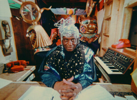 a man stares at the camera wearing an elaborate and reflective headpiece, surrounded by different objects including masks, instruments, telephones, and various props