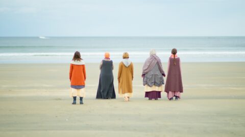 Five figures in colorful coats and hats stand on a sand beach with their backs to the camera, and the ocean in the distance.