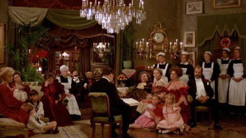 A still from the film of a well-dressed family, guests, and costumed waitstaff sitting around listening to a man read a book in an opulent interior.