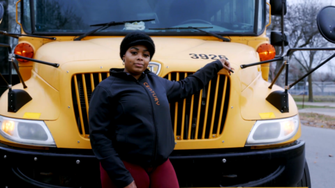 An image of a femme presenting dark skinned woman in a black shirt and hat posing in front of a school bus.