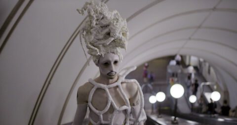 A mysterious image of a white interior with white lights and an otherworldly white costumed figure with a sculptural white hat and white faceprint with dramatic black lip liner.