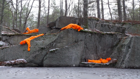 Four figures dressed and masked in safety orange lie on rock outcrops besides a body of water with snow on the ground.