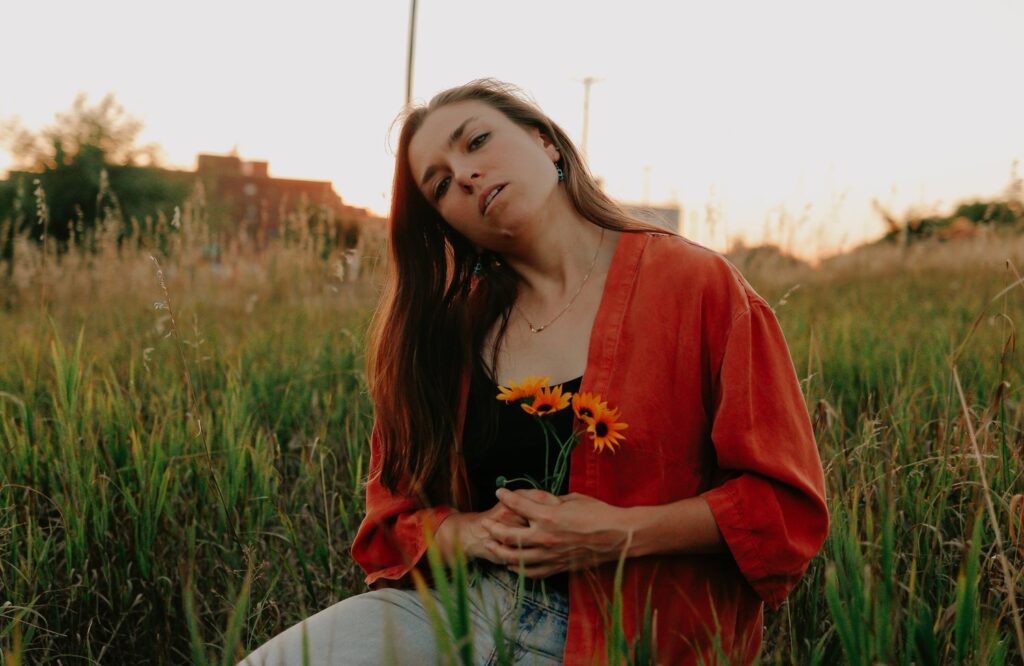 Eliza Edens in a red jacket and black shirt holding orange flowers with a setting sun glowing over the field behind her. 