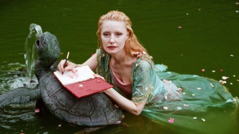 a person wading in green water resting on a turtle statue, writing in a red journal with rose petals scattered across the water