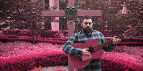 Ceschi Ramos with guitar in a field that glows with a rose-tinted hue. A cross behind him signals a graveyard? We can't be sure.