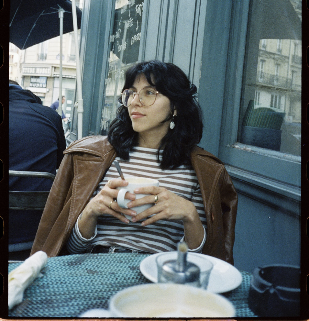 G Gilbet, a nonbinary person with long black hair, sits at a cafe table holding a mug