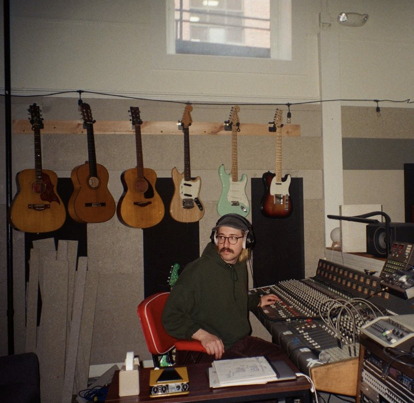 Joe Sasso (he/they) sits at a sound engineering booth with several guitars hanging on the wall behind them.