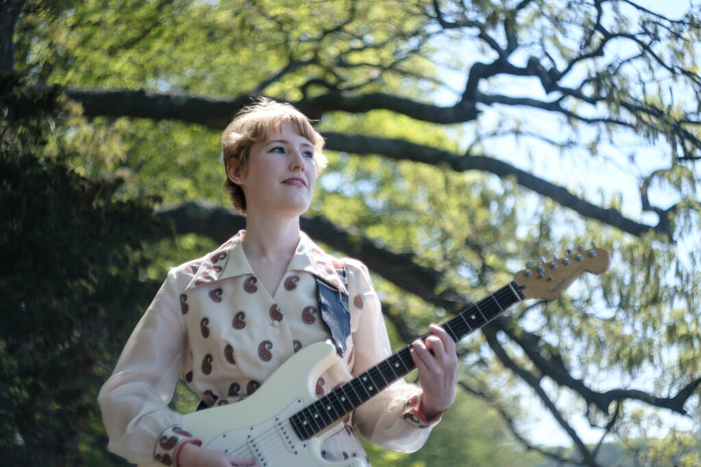 Kioea's Carand Burnet on a clear, sunny day, standing under a tree with cream-colored guitar in hand. 